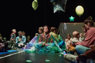 Scene from the opera for very young children Everything That Flies. At the center of the performance area, three performers sit wrapped in shimmering translucent fabrics in shades of blue, green, and purple. Toddlers and their caregivers watch from close by, seated around the stage. Balloons of different shapes float above the scene, while the open seating arrangement creates an intimate and interactive atmosphere.