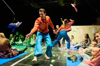 Scene from the opera for very young children Everything That Flies. Two female performers and one male performer move among toddlers and their caregivers, who are seated on mats around the performance area. Colorful bird-shaped objects hang above the stage, while feathers are scattered across the floor. The performance creates an open and interactive atmosphere, allowing the young audience to experience the action up close.