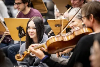A group of participants in the Crashkurs Oper sits among the musicians of the orchestra during a rehearsal. In the center, a young woman with long dark hair and a grey hoodie looks directly at the camera while seated between the instrumentalists. Around her, orchestra members play violins and woodwind instruments, with music stands and sheet music visible throughout the room. The close proximity between audience participants and musicians conveys an immersive and personal insight into the world of opera and