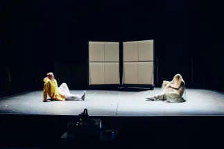 A sparsely lit stage shows two people sitting opposite each other. On the left, one person wears a yellow raincoat over white clothing; on the right, another person in light-coloured clothing sits beneath a net-like fabric. Two bright rectangular stage elements stand in the background. The scene appears focused and experimental. Teenagers and adults explore expression, movement and theatrical performance in a hands-on opera workshop.