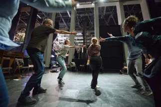 Several teenagers and adults move towards one another with outstretched arms in a large rehearsal space. The participants stand in a circle and explore shared performance situations through movement, gesture and interaction. Chairs, stage equipment and tall structural supports can be seen in the background. The scene conveys energy, concentration and creative collaboration in a hands-on opera workshop.