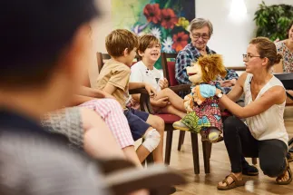 In a community room, children from a daycare centre sit together with senior citizens in a circle of chairs. A workshop leader kneels in front of the participants and guides the encounter using a colourful hand puppet. Two children respond attentively and joyfully to the puppet, while an older woman watches with interest. The warm atmosphere and lively interaction between the generations convey exchange, connection, and shared experiences.