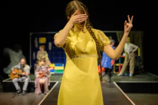 A young person in a yellow dress stands at the front of a stage. With one hand they cover their eyes, while the other forms an expressive gesture. In the background, several other participants can be seen, along with two seated musicians playing guitars. The stage is simply designed and illuminated by warm lighting. The scene conveys self-expression, self-discovery, and the emergence of one's own identity.