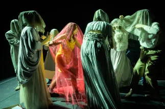 On a dark stage, a group of teenagers and adults stands closely together. Several people are covered with light, translucent fabrics, while one person emerges at the centre beneath a bright red veil. The participants move towards one another, creating an expressive scene that explores the tension between concealment and visibility. The image shows creative stage work in the hands-on opera workshop “Don’t Hide Yourself.”