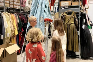 The “OPERators” of Oper Köln visit the costume department. Several children stand among densely packed clothing racks and historic stage costumes while a staff member explains the costume collection to them. In the background, elaborately designed garments from different periods and in various colours are hanging. The scene offers a glimpse into the behind-the-scenes work of the opera house.