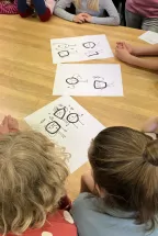 Several children are sitting in a circle on the floor, looking together at drawings by Tim Stadie. The sheets show simple characters representing the “OPERators” of Oper Köln. The children are discussing the figures and developing them further. The scene conveys focused and creative group work.