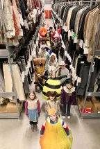 The “OPERators” of Oper Köln proudly present themselves in a variety of imaginative opera costumes. The children stand in an aisle between tall shelves in the costume department, wearing colourful, historical, and unusual outfits, including a bee, a flower, a knight, a chocolate bar, and characters from different eras. The group looks proudly at the camera, showcasing the diversity of costumes from Oper Köln’s extensive costume collection.