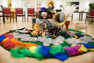 Intergenerational music theatre project Unisono: In a bright room, a colourful circle of scarves, instruments, puppets, and everyday objects is arranged on the floor. This creative landscape of play and sound invites people of different generations to experience music, stories, and meaningful encounters together.