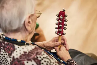 An older woman attends an Oper.Stadt.Schule event and holds a handheld instrument with numerous small bells. The photograph is taken from behind, focusing on her hands and the instrument. Her calm, attentive posture conveys active music-making, participation, and the joy of experiencing music together.