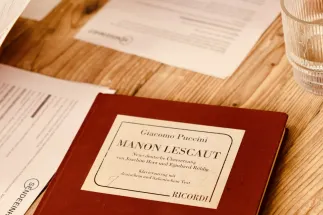 A red vocal score of Giacomo Puccini’s Manon Lescaut lies on a wooden table. Beside it are notes, printed documents, and a glass of water. The close-up photograph conveys the focused working atmosphere of a podcast recording session and shows materials used in the preparation and discussion of an opera production.