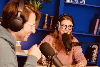 The two podcast hosts, Anna Mangold and Sebastian Sonntag, sit in a recording studio wearing headphones and speaking into microphones. They are smiling and engaged in conversation. Books and plants on a blue shelving unit can be seen in the background. The photograph conveys a focused yet relaxed atmosphere during the podcast recording.