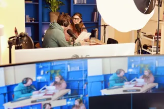 The two podcast hosts, Anna Mangold and Sebastian Sonntag, sit at a table wearing headphones and speaking into microphones. The photograph shows the studio from the control room’s perspective: monitors displaying the live recording are visible in the foreground, while lighting equipment, microphones, and technical gear reveal the professional production environment.