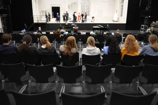 Mehrere Personen sitzen im Zuschauerraum und verfolgen eine Opernprobe auf der Bühne. Während Sängerinnen und Sänger in einem Bühnenbild proben, beobachten die Teilnehmenden aufmerksam das Geschehen. Die Aufnahme vermittelt einen Einblick in die Vorbereitung des Podcasts und die begleitende Recherche bei einem Probenbesuch.