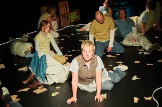 On a stage, several teenagers and young adults are sitting and lying in an expressive group scene. In the foreground, a person with short blond hair supports themselves on their hands while looking intently ahead. Around them, the other performers are spread across the dark stage floor, which is covered with torn pieces of paper. The scene is from the performance When I Find Myself in Times of Trouble by the Opera, Song and Scene performance club.
