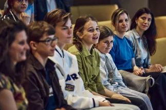 Members of the OPERators of Opera Cologne take part in the Children's Opera Cologne press conference for the 2026/27 season. Several children and teenagers are seated attentively in a discussion round, listening with focus. Some look ahead with interest, while others follow the conversation with curious expressions. The image conveys the young experts' active participation in the exchange about the Children's Opera programme.