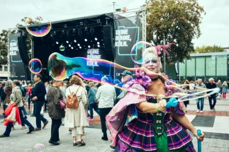 Ein Mitglied des Chors der Oper Köln in einem bunten Kostüm erzeugt Seifenblasen beim Eröffnungsfest der Bühnen Köln. Im Hintergrund stehen Besucher*innen vor der Bühne.
