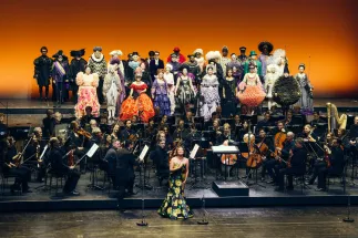Adriana Bastidas-Gamboa steht singend vor dem Gürzenich-Orchester Köln. GMD Andrés Orozco-Estrada dirigiert; dahinter singt der Chor der Oper Köln in farbenprächtigen Kostümen.
