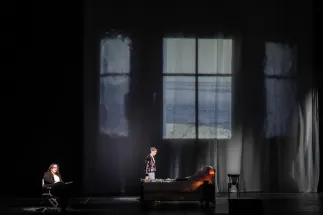 The Marschallin, Princess Werdenberg (Magdalena Hinterdobler), sits on the left with a book. Octavian (Bianca Andrew) stands beside a bed on the right. In the background, a projection shows a view of the sea through a window. A scene from Richard Strauss’s “Der Rosenkavalier”.