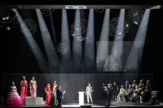 A scene from Richard Strauss’s “Der Rosenkavalier” featuring Sophie (Giulia Montanari), Octavian (Bianca Andrew), the three noble orphans (Kristi Anna Isene, Jing Yang and Martina Sigl), Marianne Leitmetzerin (Claudia Rohrbach), the police commissioner/notary (Lucas Singer), Faninal’s steward (Wesley Harrison), a singer (Dmitry Ivanchey) and the Cologne Opera Chorus. The performers stand and sit in groups on platforms. Beams of light fall across the stage, with illuminated roses hanging above.