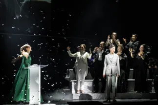 Sophie (Giulia Montanari) stands on the left in a green dress with her arms outstretched, while Octavian (Bianca Andrew) stands on the right in a light grey suit. Members of the Cologne Opera Chorus applaud on a platform in the background. Confetti falls across the stage. A scene from Richard Strauss’s “Der Rosenkavalier”.