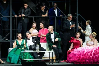 A festive scene from Richard Strauss’s “DER ROSENKAVALIER”: Sophie (Giulia Montanari), wearing a green dress, sits on the left beside Baron Ochs of Lerchenau (Patrick Zielke). Around them, Herr von Faninal (Christian Miedl), Marianne Leitmetzerin (Claudia Rohrbach), the three noble orphans (Kristi Anna Isene, Jing Yang and Martina Sigl) and members of the Cologne Opera Chorus stand and sit. Some hold glasses of sparkling wine.