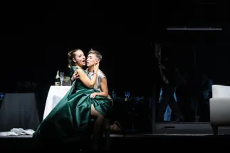 Sophie (Giulia Montanari), wearing a green dress, and Octavian (Bianca Andrew), wearing a light grey suit, sit in a close embrace on the dark stage. Behind them is a table with glasses. A scene from Richard Strauss’s “Der Rosenkavalier”.