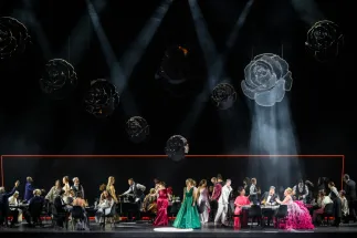 A festive gathering in Richard Strauss’s “Der Rosenkavalier”: Numerous people sit and stand around tables on stage. In the foreground, a green dress and a voluminous pink gown stand out. Large rose shapes hang above them, illuminated by beams of light.