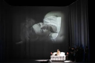 On stage, the Marschallin, Princess Werdenberg (Magdalena Hinterdobler), and Octavian (Bianca Andrew) sit in a bed. A live black-and-white close-up of the Marschallin is projected above them. A scene from Richard Strauss’s “Der Rosenkavalier”.