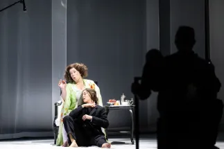 The Marschallin, Princess Werdenberg (Magdalena Hinterdobler), sits on a chair and gestures while Octavian (Bianca Andrew) leans against her on the floor. Dark silhouettes obscure part of the stage on the right. A scene from Richard Strauss’s “Der Rosenkavalier”.