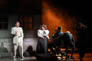 A singer (Young Woo Kim) stands on the left holding a hat, with the Marschallin, Princess Werdenberg (Magdalena Hinterdobler), beside him. On the right, members of the Cologne Opera’s extra cast operate a camera and hold a microphone above the scene. Orange light falls across the stage. A scene from Richard Strauss’s “Der Rosenkavalier”.