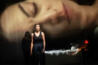 The Marschallin, Princess Werdenberg (Magdalena Hinterdobler), stands in front of an unmade bed. Octavian (Bianca Andrew) stands in the background on the left, with her back to the camera. An oversized image of Octavian’s face is projected onto the stage wall. A scene from Richard Strauss’s “Der Rosenkavalier”.
