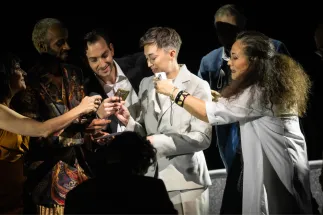 Octavian (Bianca Andrew) stands in a light-coloured suit surrounded by Cologne Opera’s extras. Several people hold objects out to her; Annina (Kammersängerin Dalia Schaechter) stands to her right. A scene from Richard Strauss’s “Der Rosenkavalier”.