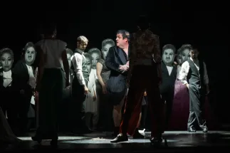 Baron Ochs of Lerchenau (Patrick Zielke), wearing a bathrobe, stands among members of the Cologne Opera’s extra cast on a dark stage. Some hold oversized portraits in front of their faces. A scene from Richard Strauss’s “Der Rosenkavalier”.