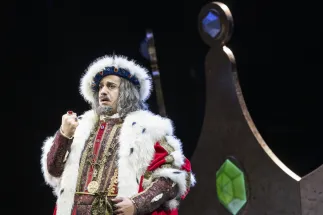 The old King Detlef (Ferhat Baday) raises his ring-adorned fist. He wears a red and gold robe trimmed with white fur, a crown, and a grey wig with a beard. Scene from the children’s opera “DER KÖNIG VERSCHENKT WAS” by Philipp Matthias Kaufmann.