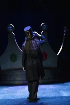 Photographer / Servant / Death (Anna Kelly) stands with her back to the audience in front of a large crown, wearing a black coat with bone motifs and a top hat with a glowing skull. Scene from Philipp Matthias Kaufmann’s DER KÖNIG VERSCHENKT WAS.