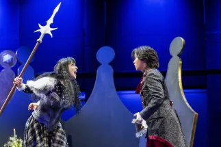 Jeannette (Laura Kriese) and Louis (Geonmuk Lim) face each other in front of a large crown. Jeannette wears a breastplate and holds a lance. Louis wears a black, patterned, glittering coat with red embellishments. Scene from Philipp Matthias Kaufmann’s DER KÖNIG VERSCHENKT WAS.