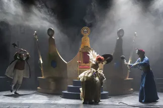 King Friedrich from the West (Wonjun Kim), Queen Elisabeth from the South (Lena Geiger) and King Günther from the East (Tobias Lusser) stand with weapons in front of a large golden crown and a throne. Friedrich wears a green coat and raises an axe above his head. Elisabeth wears a golden dress with a high white collar and holds a black shield. Günther wears a blue robe and swings a flail. Scene from Philipp Matthias Kaufmann’s DER KÖNIG VERSCHENKT WAS.