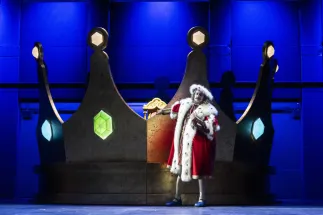 The old King Detlef (Ferhat Baday) stands in front of a large crown with glowing gemstones. He wears a red royal robe trimmed with white, black-spotted fur and a matching headdress. One arm rests on the crown, while he holds his other hand in front of his chest. Scene from Philipp Matthias Kaufmann’s DER KÖNIG VERSCHENKT WAS.