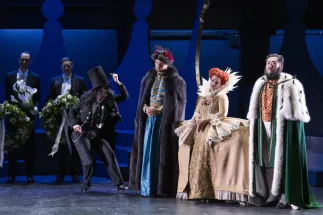 Two extras from Cologne Opera hold floral wreaths. Photographer / Servant / Death (Anna Kelly) wears a black suit, sunglasses and a top hat and raises one arm. Beside her stand King Günther from the East (Tobias Lusser) in a blue robe with a dark fur coat, Queen Elisabeth from the South (Lena Geiger) in a golden dress with a high white collar and King Friedrich from the West (Wonjun Kim) in a green coat trimmed with white fur. Funeral scene from Philipp Matthias Kaufmann’s DER KÖNIG VERSCHENKT WAS.