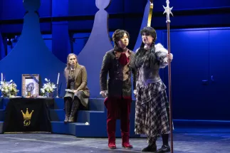 Claire (Lana Sophie Westendorf), wearing a brown blazer, sits on a step beside a memorial table decorated with flowers, candles and a portrait of the king. In front stand Louis (Geonmuk Lim) in a black glittering coat with red embellishments and Jeannette (Laura Kriese) wearing a breastplate and a checked skirt and holding a lance. Scene from Philipp Matthias Kaufmann’s DER KÖNIG VERSCHENKT WAS.