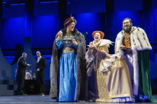 King Günther from the East (Tobias Lusser) in a blue robe with a dark fur coat, Queen Elisabeth from the South (Lena Geiger) in a golden dress with a high white collar and King Friedrich from the West (Wonjun Kim) in a green coat trimmed with white fur stand side by side, laughing and holding plates full of food. In the background, two extras from Cologne Opera stand beside a memorial table with a portrait of the king. Scene from Philipp Matthias Kaufmann’s DER KÖNIG VERSCHENKT WAS.