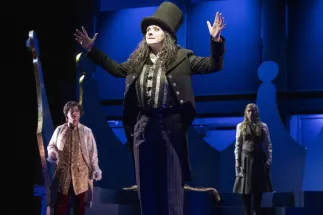 Photographer / Servant / Death (Anna Kelly) stands in the foreground with her arms raised, wearing a black tailcoat, a silver-striped waistcoat and a tall top hat. Behind her stand Louis (Geonmuk Lim) in a light-coloured coat over a golden garment and Claire (Lana Sophie Westendorf) in a white blouse, a beige-brown waistcoat and a grey skirt. Scene from Philipp Matthias Kaufmann’s DER KÖNIG VERSCHENKT WAS.