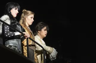 Jeannette (Laura Kriese), Claire (Lana Sophie Westendorf) and Louis (Geonmuk Lim) stand side by side at a black railing, looking to the right. Jeannette wears a breastplate with white fur trim and a checked skirt, Claire a golden coat over a beige-brown waistcoat and Louis a cream-coloured, patterned coat with lace cuffs. Scene from Philipp Matthias Kaufmann’s DER KÖNIG VERSCHENKT WAS.