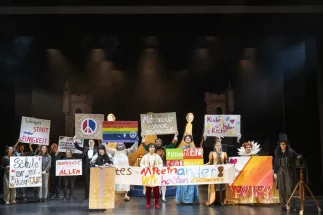 The performers stand side by side on stage, holding colourful, handmade posters about peace, democracy, human rights and solidarity. A long banner stretches across the centre, reading “Together! We stick together”. Scene from Philipp Matthias Kaufmann’s DER KÖNIG VERSCHENKT WAS.