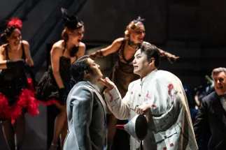 Rigoletto (Insik Choi) hält eine Hand unter das Kinn von Graf Ceprano (Michael Terada), der einen grauen Anzug trägt. Rigoletto trägt ein weites, helles Kostüm mit bunten Mustern und hält einen spitzen Hut. Hinter ihnen stehen lachende Tänzerinnen in Abendkleidern mit Federbesatz und Kopfschmuck; rechts ist Borsa (Yves Saelens) im dunklen Anzug zu sehen. Szene aus Giuseppe Verdis RIGOLETTO.