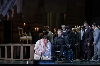 Rigoletto (Insik Choi) kauert im hellen, bunt gemusterten Kostüm auf dem Bühnenboden und hält sich eine Hand an den Hals. Neben ihm sitzt Graf von Monterone (Christoph Seidl) im dunklen Anzug in einem Rollstuhl. Dahinter stehen Borsa (Yves Saelens), Marullo (Tobias Lusser), Graf Ceprano (Michael Terada) und der Herrenchor der Oper Köln. Kerzenleuchter erhellen die dunkle Bühne. Szene aus Giuseppe Verdis RIGOLETTO.