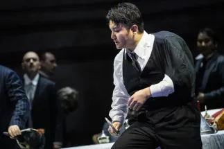 Rigoletto (Insik Choi) sitzt nach vorn gebeugt auf einer Tischkante und blickt nach unten. Er trägt ein weißes Hemd, eine schwarze Weste, Krawatte und Hose; über seinem Rücken liegt ein dunkles Jackett. In einer Hand hält er einen kleinen Spiegel. Im Hintergrund stehen Männer in dunklen Anzügen. Szene aus Giuseppe Verdis RIGOLETTO.