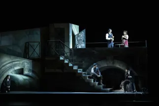 Gilda (Enkeleda Kamani) kauert links unter einem steinernen Bogen. Sparafucile (William Thomas) sitzt mit dunkler Mütze und Stiefeln auf einer Treppe, während Rigoletto (Insik Choi) rechts auf einem Fass sitzt und einen Hut hält. Auf der erhöhten Terrasse stehen der Herzog von Mantua (Ioan Hotea) in weißem Hemd und dunkler Weste und Maddalena (Adriana Bastidas-Gamboa) in einem Kleid mit pinkfarbenem Tuch. Beide halten Trinkgefäße. Szene aus Giuseppe Verdis RIGOLETTO.