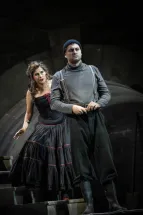 Sparafucile (William Thomas) steht in grauem Rollkragenpullover, Hosenträgern, dunkler Mütze und hohen Stiefeln auf einer Treppe. Hinter ihm steht Maddalena (Adriana Bastidas-Gamboa) in einem schwarzen Kleid mit roten Verzierungen und streckt eine Hand aus. Ein steinerner Torbogen bildet den Hintergrund. Szene aus Giuseppe Verdis RIGOLETTO.