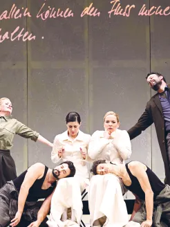 Six performers from Così fan tutte on stage: At the center sit Fiordiligi and Dorabella in elegant, cream-colored, high-necked dresses. At their feet lie Ferrando and Guglielmo, wearing dark tank tops and casual trousers, with a contemporary appearance. To the left stands Despina in an olive-green shirt and dark skirt, while Don Alfonso stands to the right in a dark coat. The costumes combine classical and modern elements.